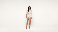 A woman stands against a plain white background, wearing a white floral blouse and matching shorts with beige high-heeled shoes. She faces forward with a neutral expression and her arms are relaxed by her sides.