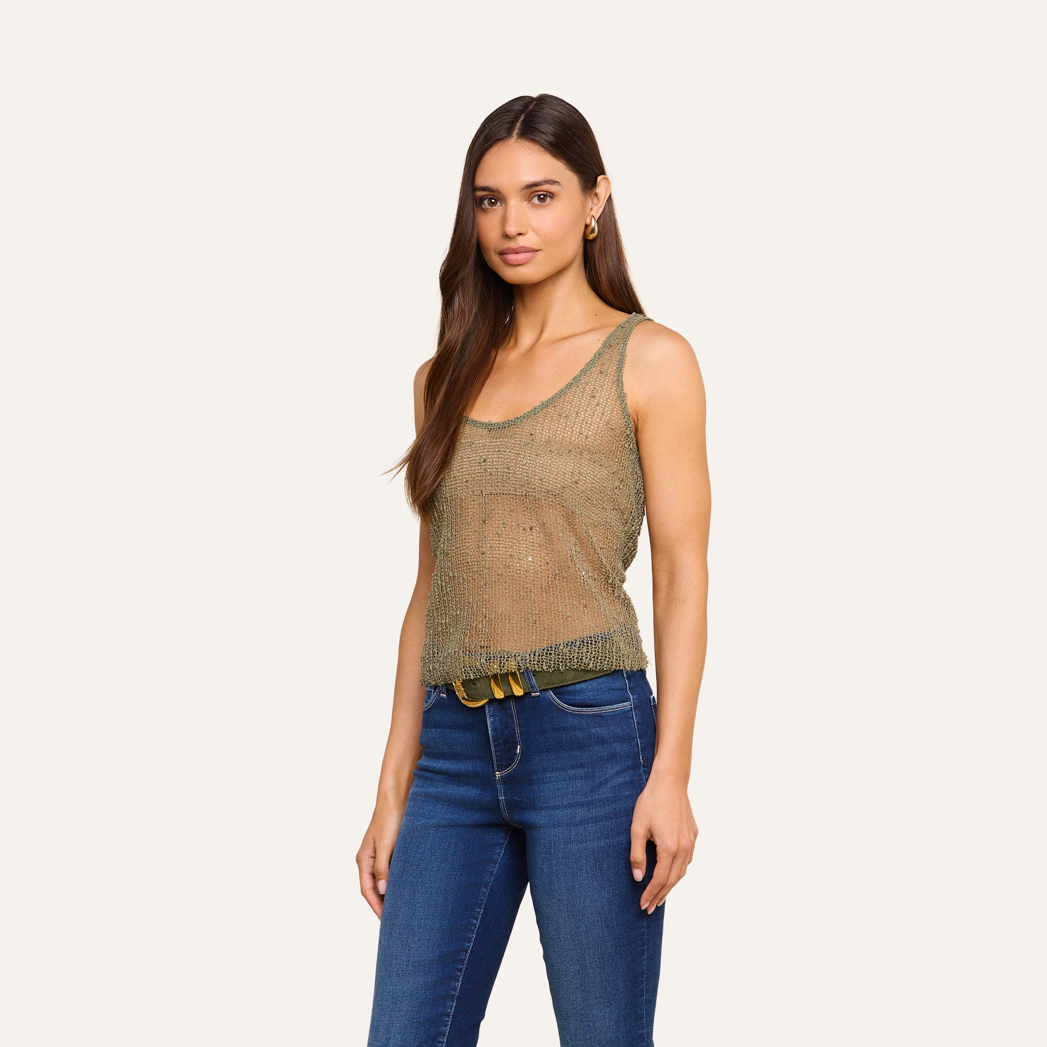 A woman with long brown hair wears the Zia Tank in olive green and blue jeans, standing against a beige background and looking at the camera with a neutral expression.