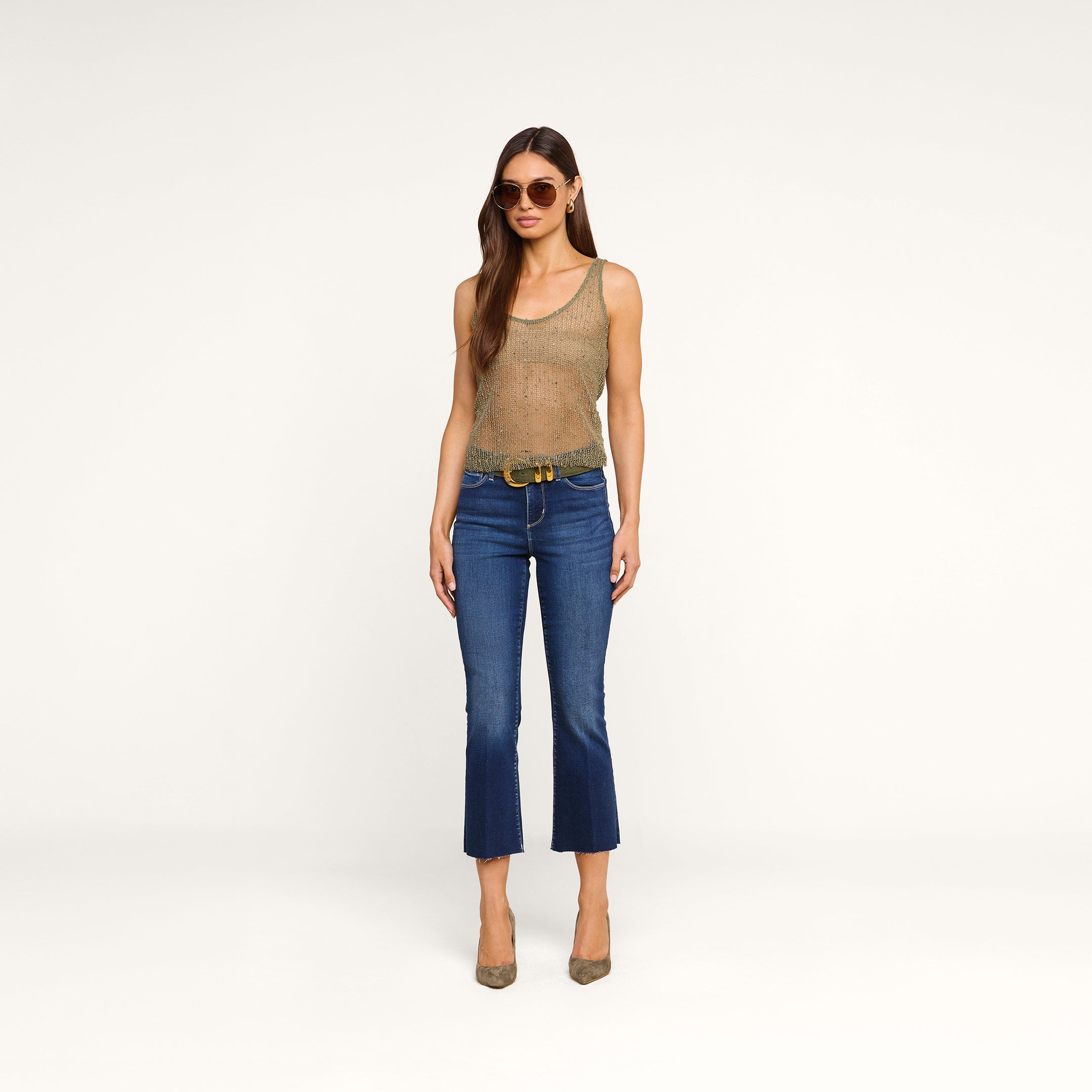 A woman with long brown hair and a confident posture stands against a beige background, wearing sunglasses, blue cropped jeans with a black belt, taupe high heels, and the Zia Tank.