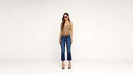 Woman wearing sunglasses, a tan tank top, blue cropped jeans, a belt, and heels stands against a plain white background, facing forward with her arms relaxed by her sides.