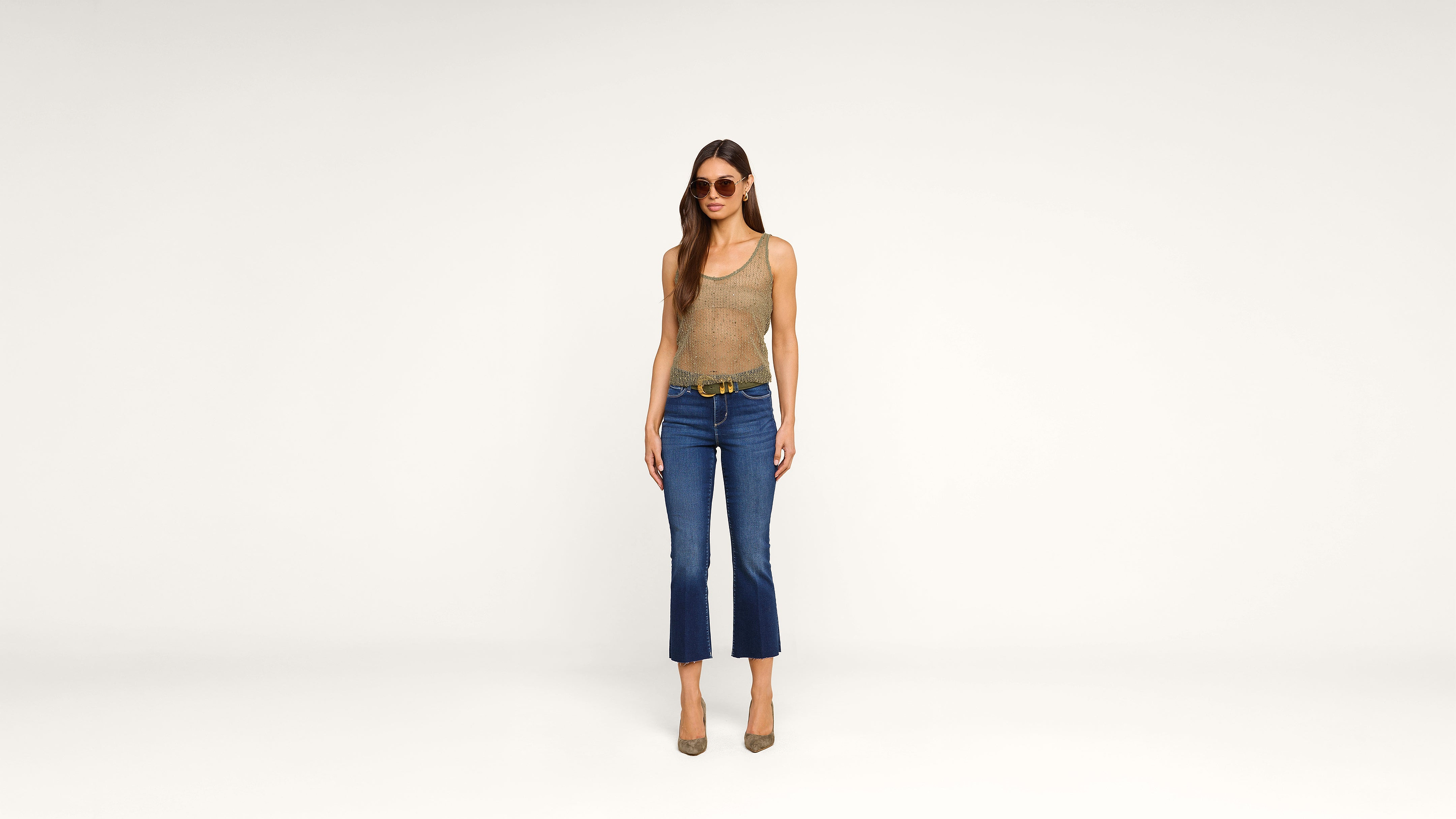 Woman wearing sunglasses, a tan tank top, blue cropped jeans, a belt, and heels stands against a plain white background, facing forward with her arms relaxed by her sides.