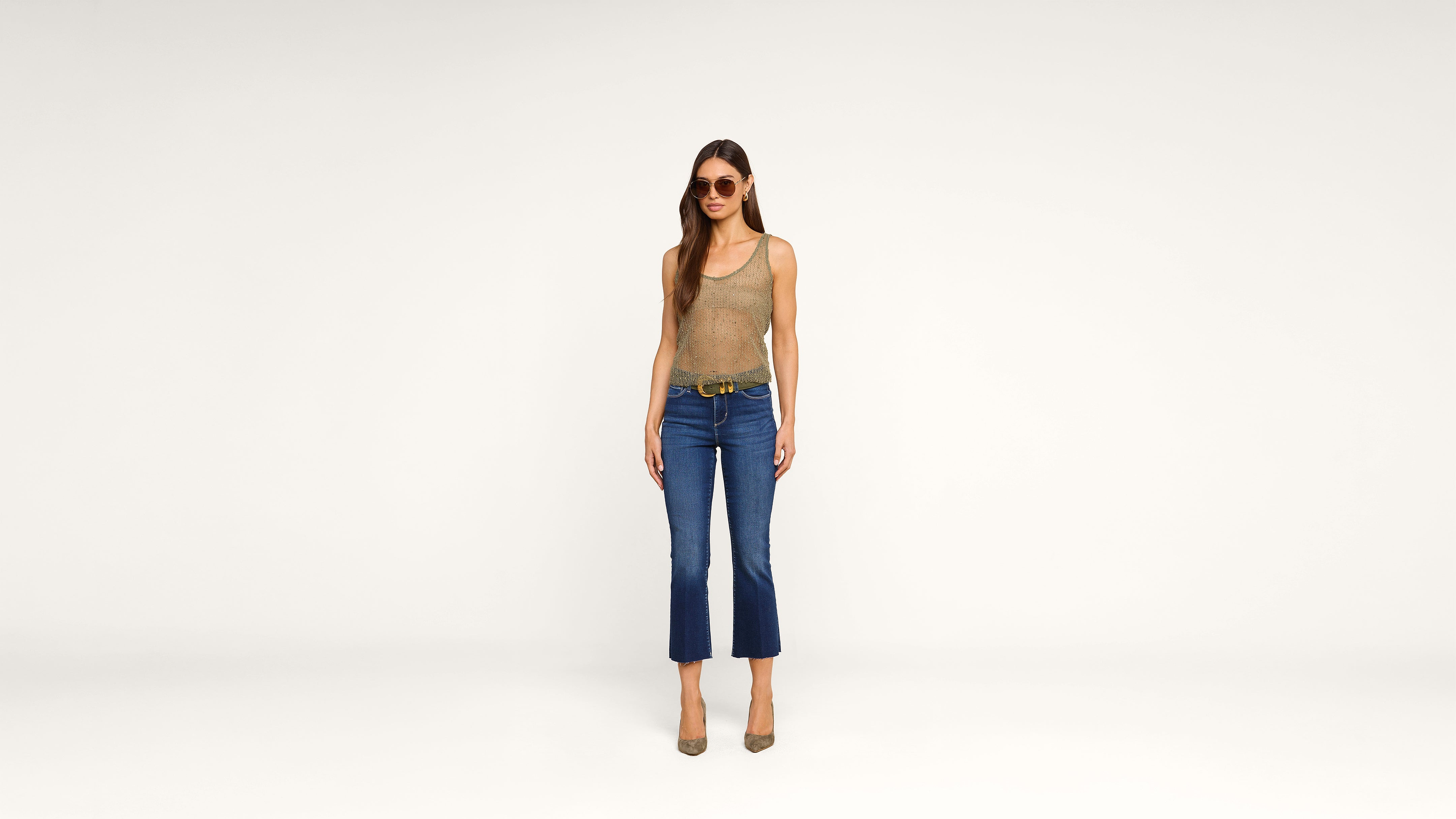 Woman wearing sunglasses, a tan tank top, blue cropped jeans, a belt, and heels stands against a plain white background, facing forward with her arms relaxed by her sides.