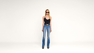 A woman wearing sunglasses, a black tank top, blue flared jeans, and black heels stands against a plain white background.