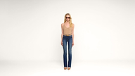 A woman wearing sunglasses, a beige sleeveless top, blue high-waisted jeans, and nude heels stands against a plain white background, facing forward.