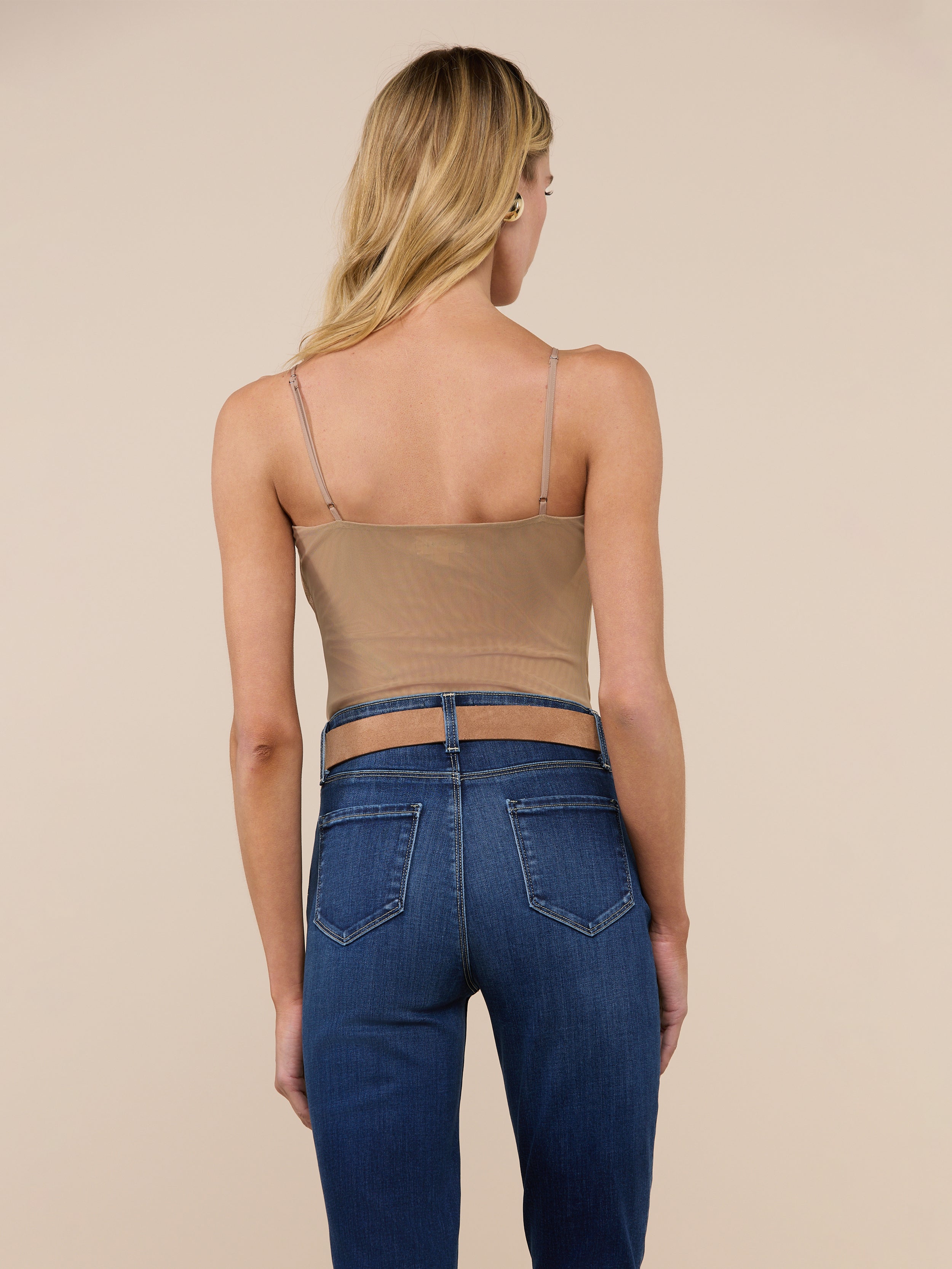 A woman with long blonde hair is shown from the back, wearing the Gelina Mesh Bodysuit—a tan spaghetti strap top with a scoop neckline—paired with blue jeans, standing against a plain, light background.