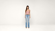 A woman stands against a plain white background wearing a pink sleeveless top, light blue high-waisted jeans, a brown belt, and nude shoes.