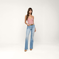 Une femme aux longs cheveux bruns pose sur un fond beige, vêtue du Bodysuit Gelina Mesh, dun jean flare bleu clair à taille haute, dune ceinture marron et de chaussures à talons pointus de couleur beige. Elle a une main dans la poche.