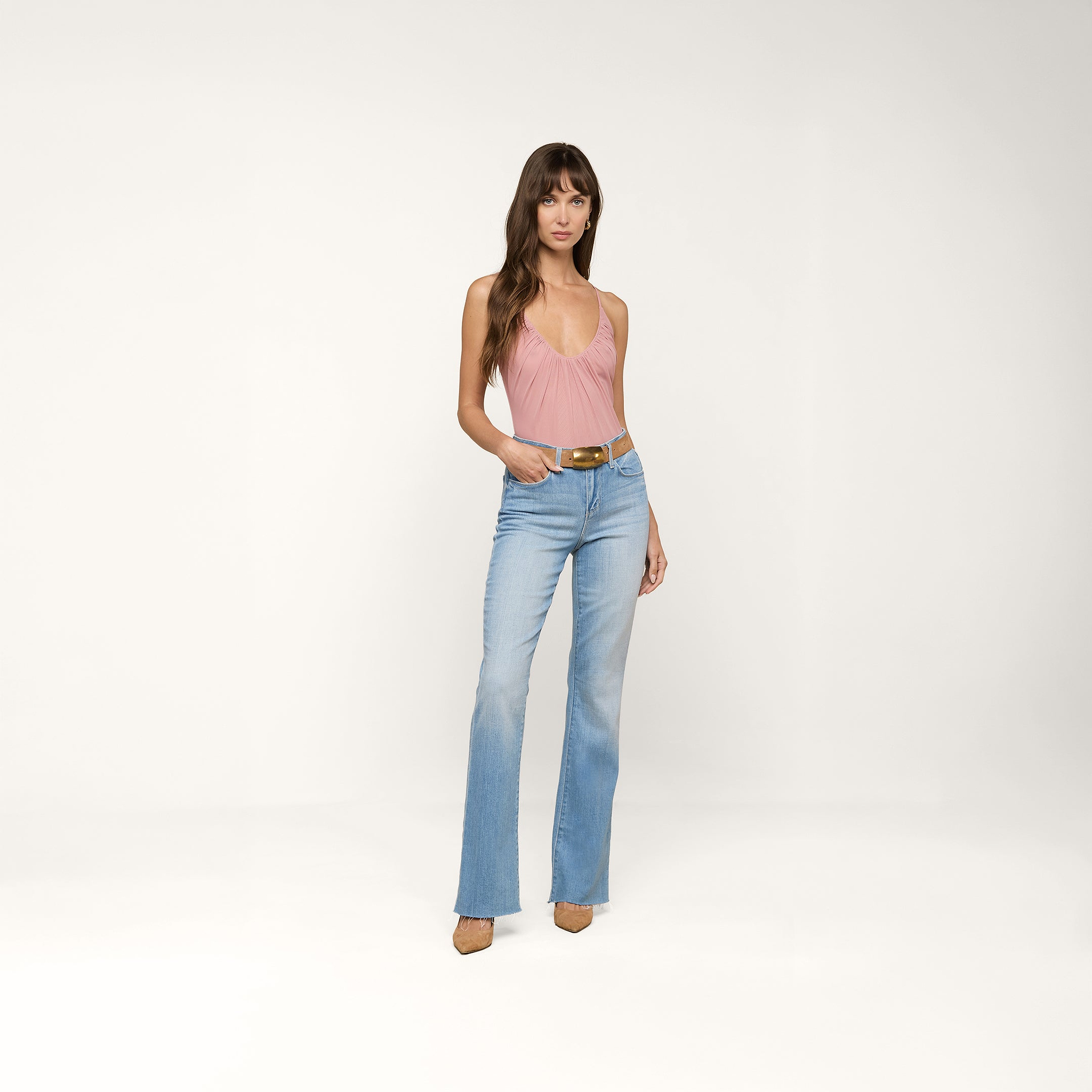 Une femme aux longs cheveux bruns pose sur un fond beige, vêtue du Bodysuit Gelina Mesh, dun jean flare bleu clair à taille haute, dune ceinture marron et de chaussures à talons pointus de couleur beige. Elle a une main dans la poche.