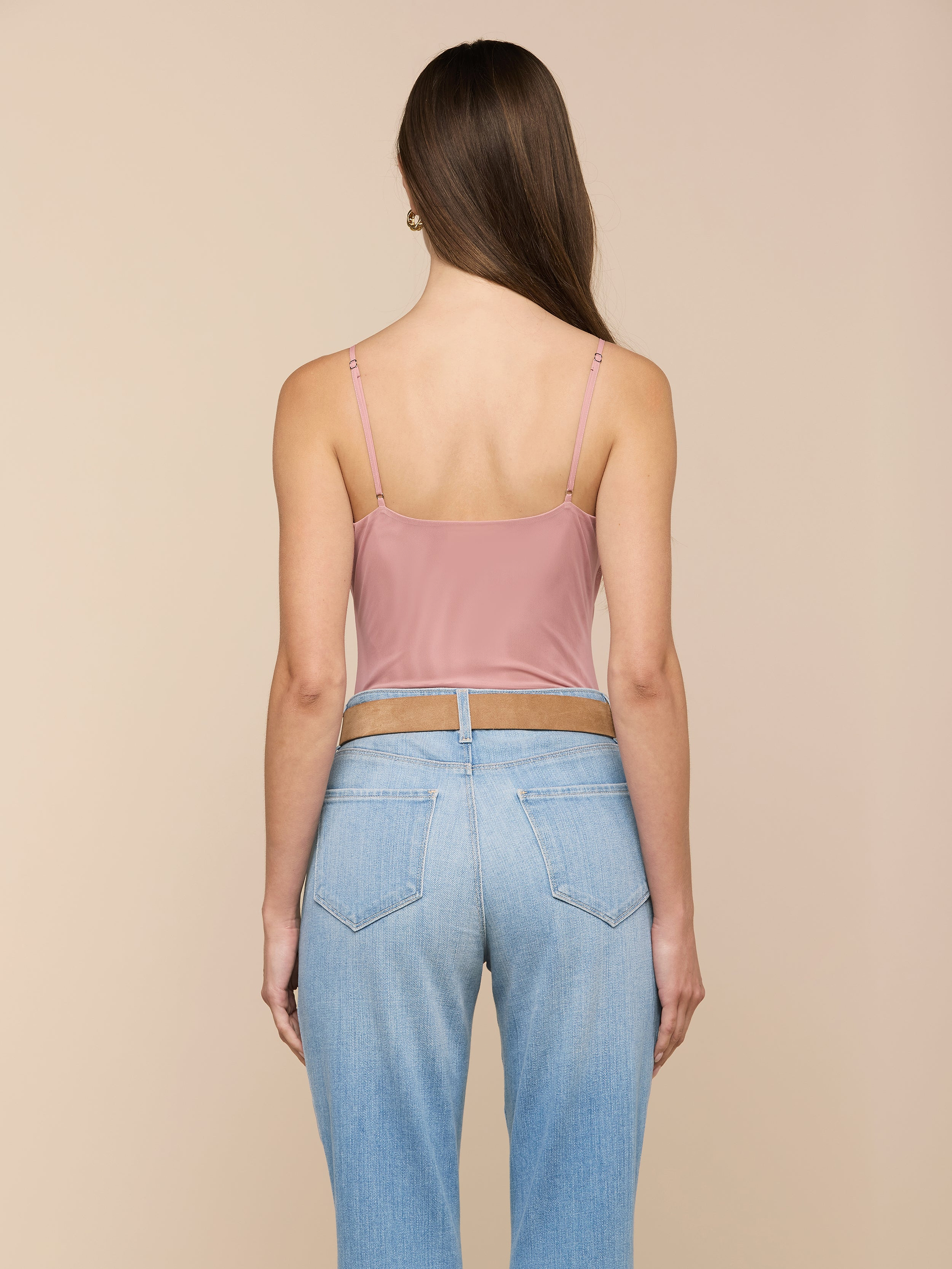 Une femme aux longs cheveux bruns se tient de face, vêtue de la combinaison Gelina Mesh Bodysuit à bretelles réglables et dun jean bleu clair à taille haute, sur fond beige.