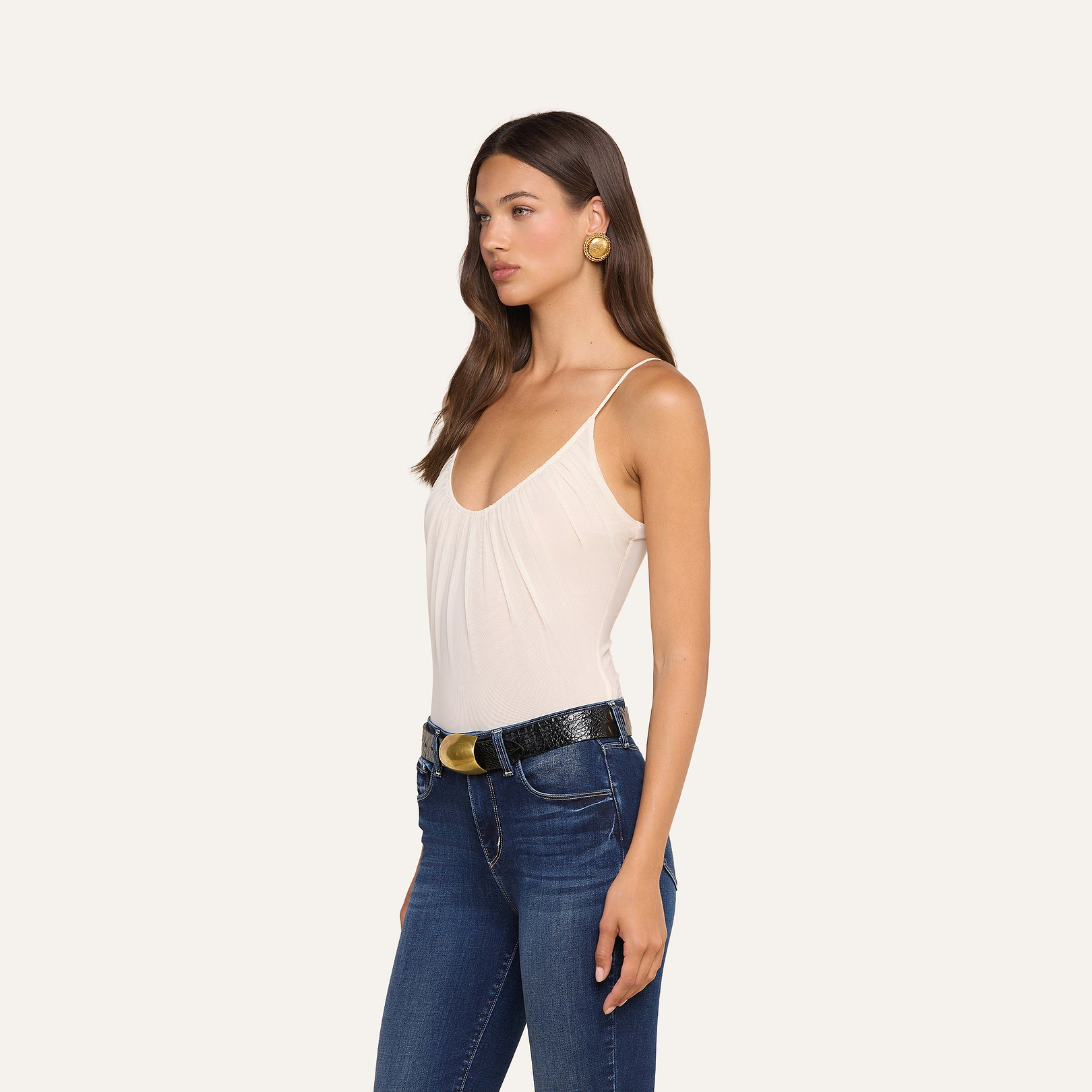 A woman with long brown hair wears gold earrings, blue jeans, a black belt with a gold buckle, and the Gelina Mesh Bodysuit—a sleeveless white top made from lightweight stretch mesh—standing against a beige background.