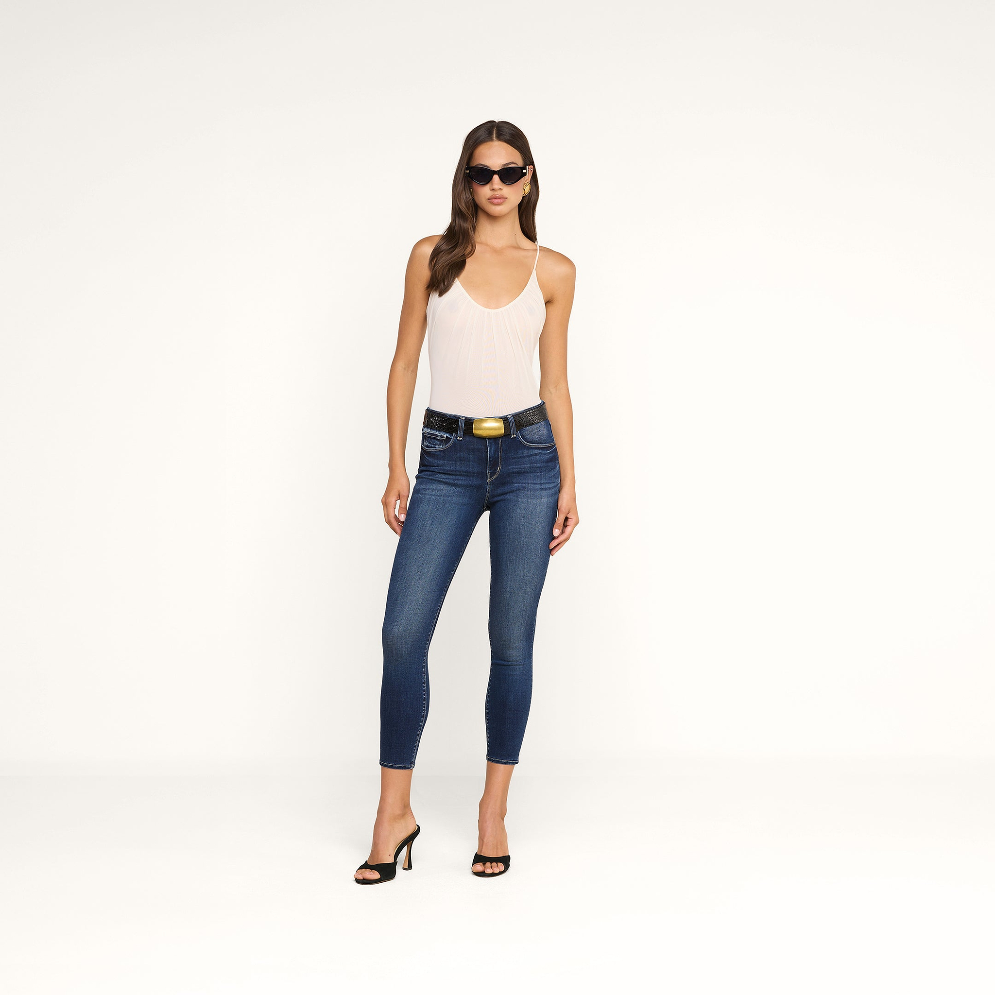 Una donna con indosso occhiali da sole, il body Gelina Mesh con spalline regolabili, jeans skinny blu con una cintura nera e sandali neri con tacco è in piedi davanti a uno sfondo beige a tinta unita.