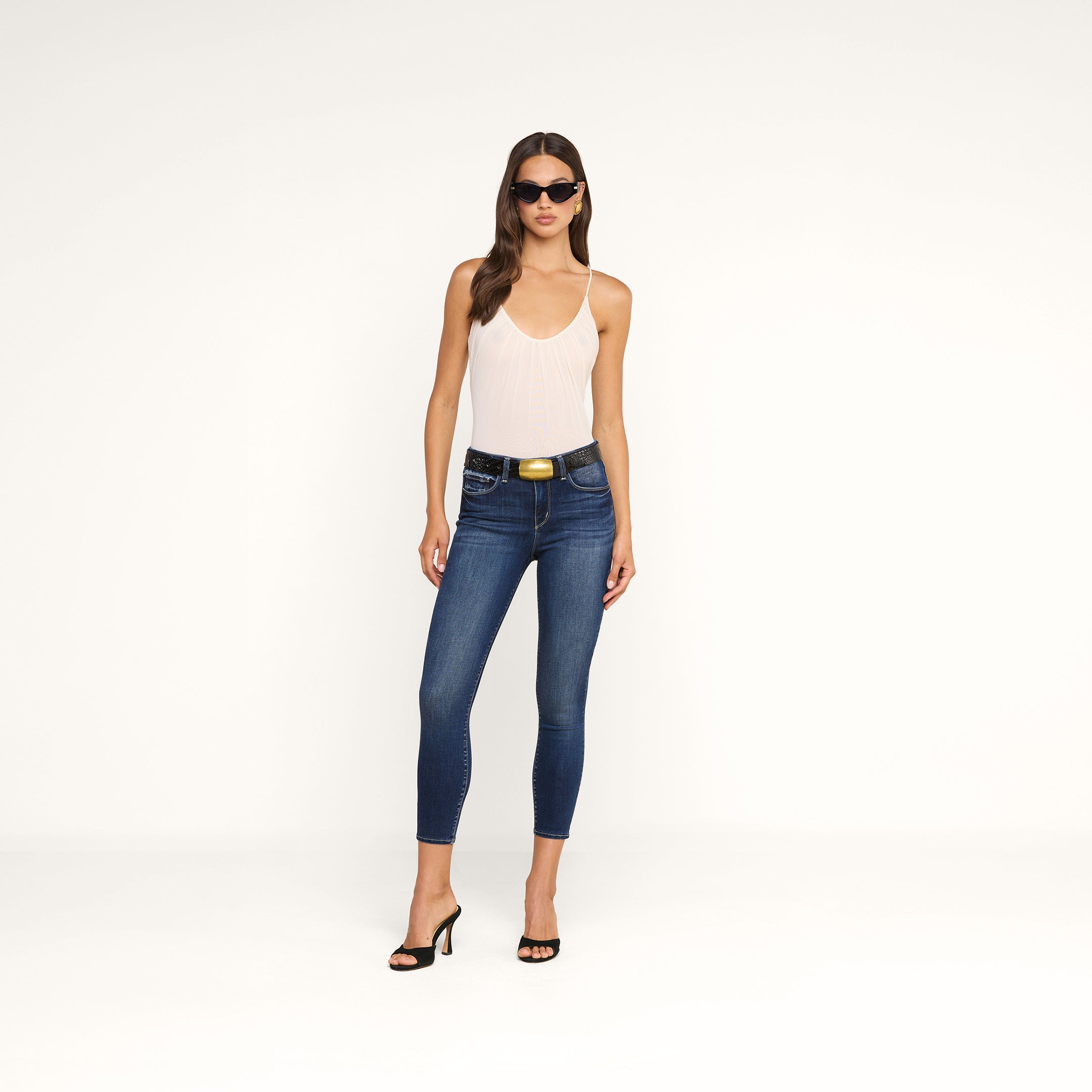 Una donna con indosso occhiali da sole, il body Gelina Mesh con spalline regolabili, jeans skinny blu con una cintura nera e sandali neri con tacco è in piedi davanti a uno sfondo beige a tinta unita.