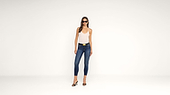 Woman wearing a white tank top, blue skinny jeans, black high-heeled sandals, and sunglasses stands against a plain white background.
