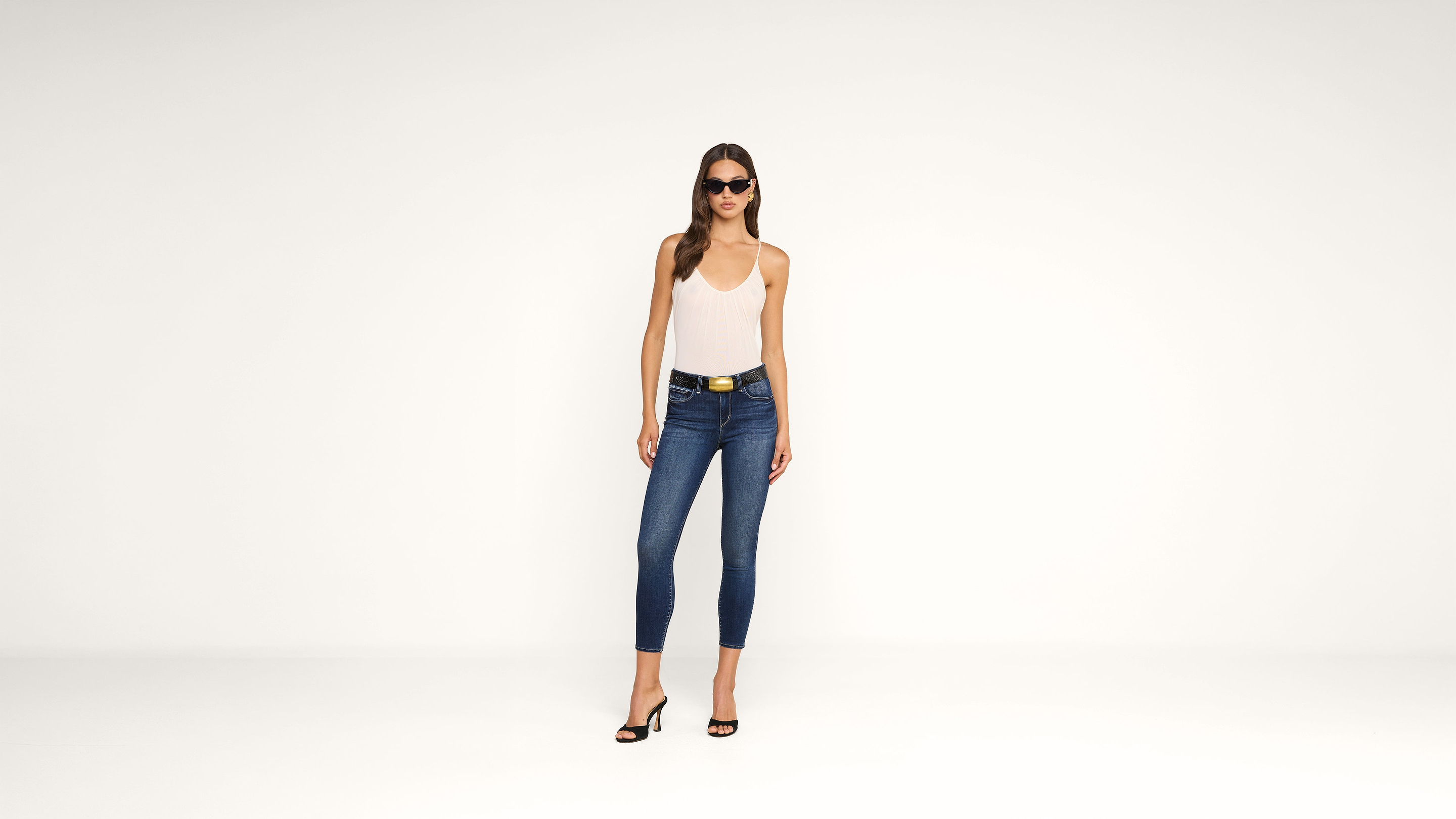 Woman wearing a white tank top, blue skinny jeans, black high-heeled sandals, and sunglasses stands against a plain white background.
