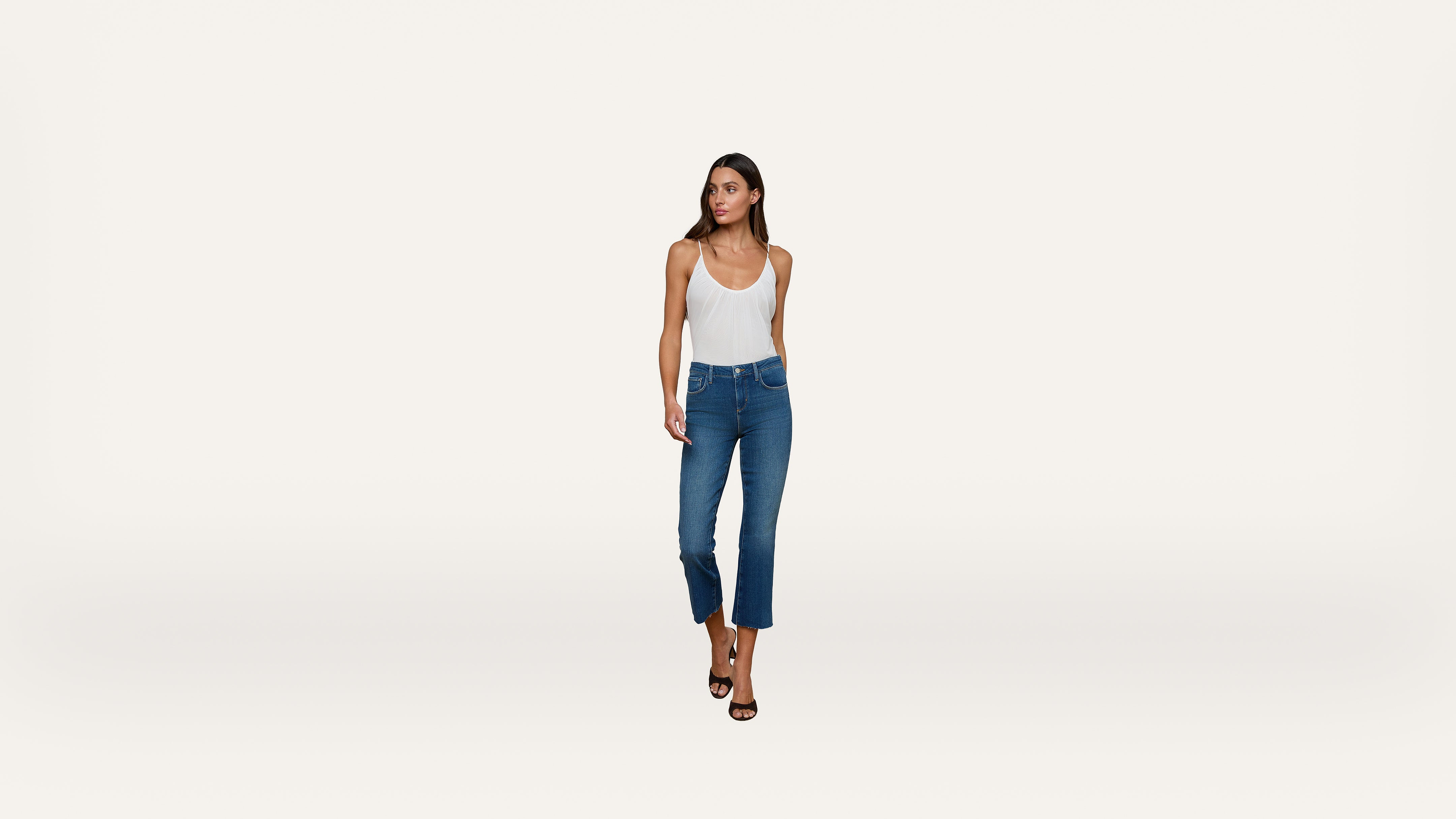A woman wearing a white tank top, blue cropped jeans, and black high-heeled sandals stands against a plain white background, looking slightly to her left.