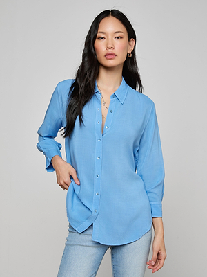 A woman with long dark hair models the light blue Marlene Blouse and matching jeans, standing against a plain background. She looks confidently at the camera with one hand in her pocket.