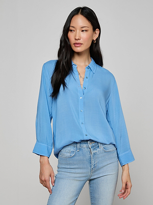 A woman with long black hair wears the Marlene Blouse—a relaxed fit, light blue, lightweight button-down with rolled sleeves—paired with light blue jeans, standing against a plain light gray background.