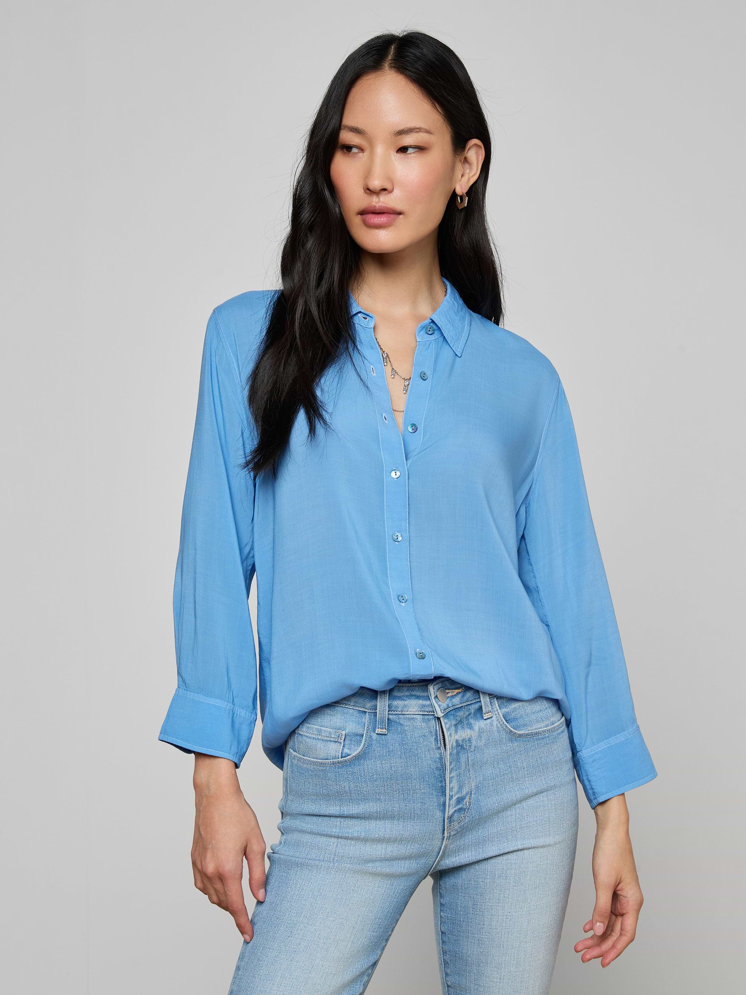 A woman with long black hair wears the Marlene Blouse, a relaxed fit, lightweight button-down shirt in light blue, paired with matching light blue jeans. She stands against a plain off-white background, looking slightly to her left with a relaxed expression.