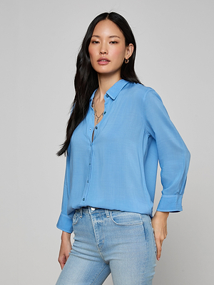 A woman with long black hair wears the Marlene Blouse in light blue and light-wash jeans, standing against a plain gray background with one hand in her pocket and a neutral expression.