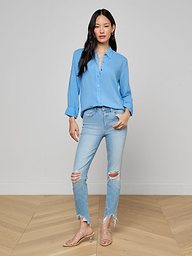 A woman wears the Marlene Blouse, a lightweight light blue button-down shirt, styled with ripped light-wash jeans and beige heels. She stands against a plain white background, facing forward with one hand in her pocket for an effortlessly relaxed fit and everyday polish.