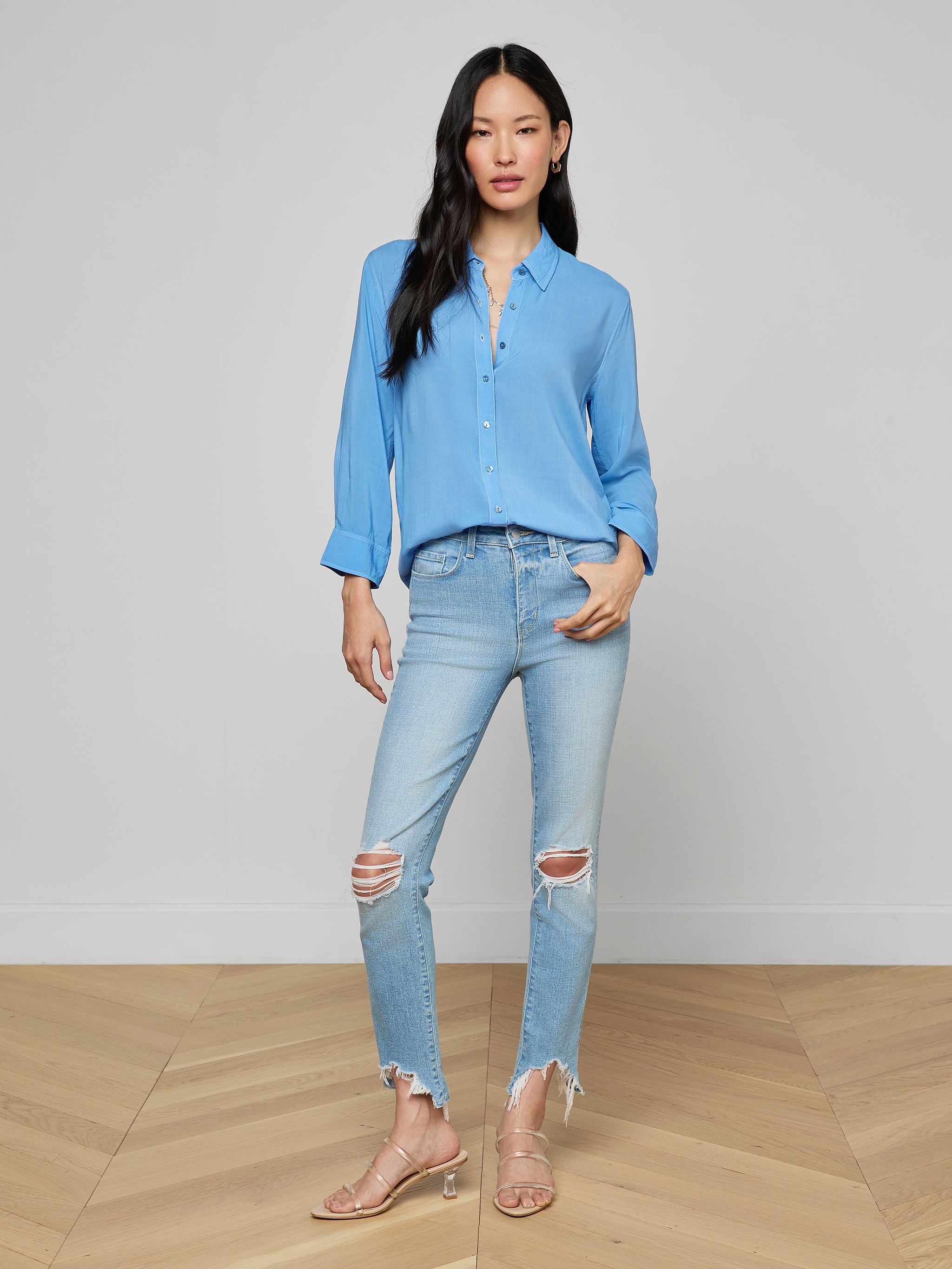 A woman wears the Marlene Blouse, a lightweight light blue button-down shirt, styled with ripped light-wash jeans and beige heels. She stands against a plain white background, facing forward with one hand in her pocket for an effortlessly relaxed fit and everyday polish.