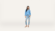Woman standing against a plain white background, wearing a light blue blouse, ripped light blue jeans, and beige sandals. She has long dark hair and her hands are in her pockets.