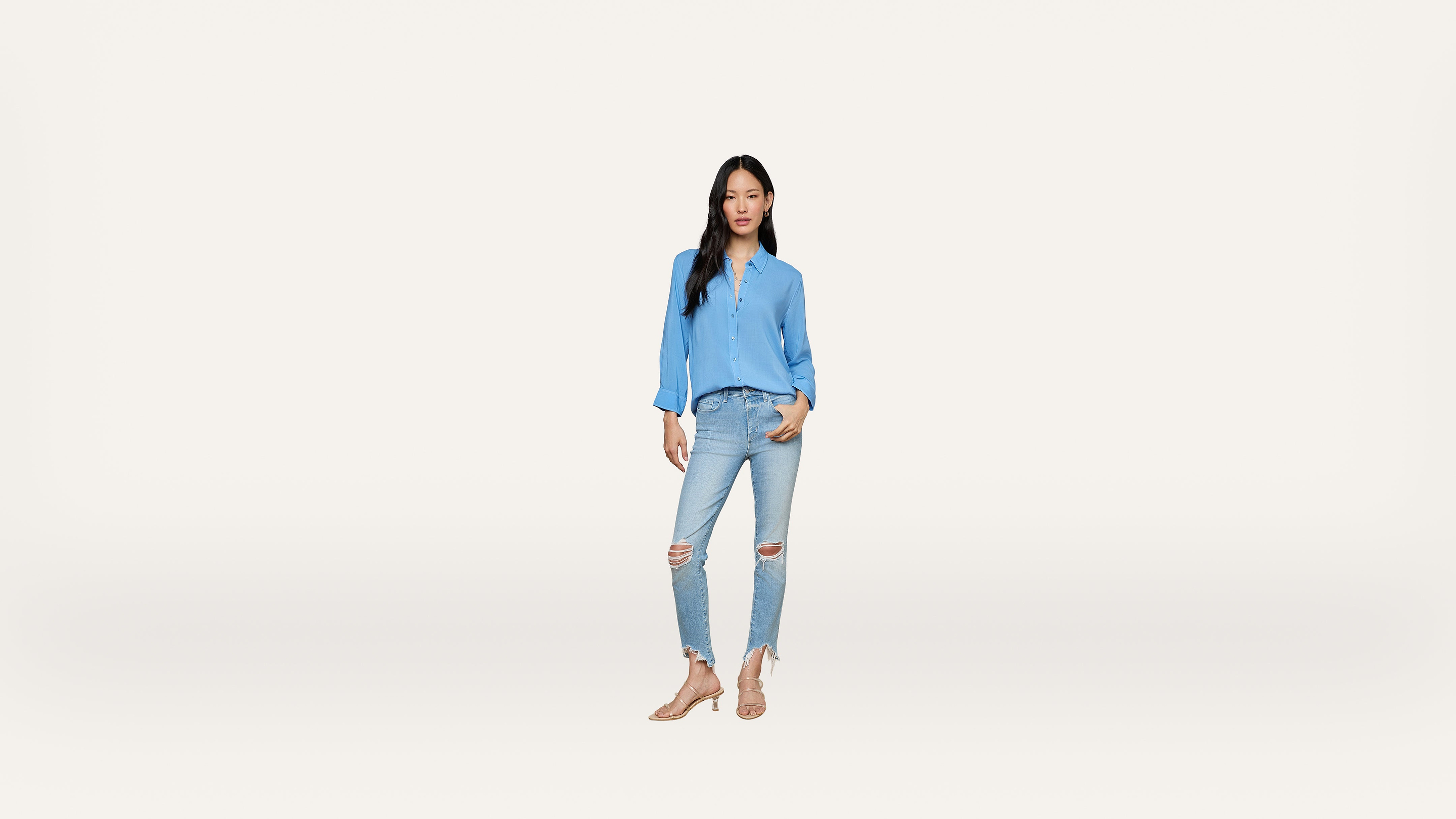 Woman standing against a plain white background, wearing a light blue blouse, ripped light blue jeans, and beige sandals. She has long dark hair and her hands are in her pockets.