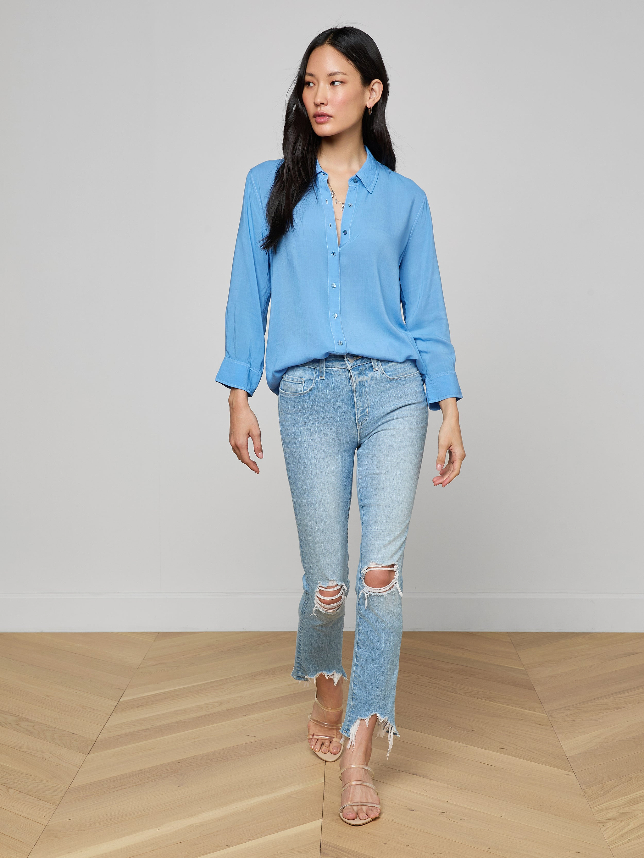 A woman wearing the Marlene Blouse, a relaxed-fit, lightweight button-down in light blue, pairs it with ripped light-wash jeans and beige sandals while standing against a plain white background for an effortless touch of everyday polish.