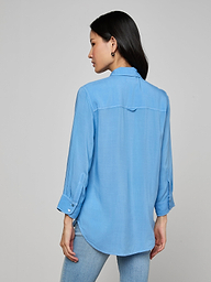 A woman stands facing away, wearing the Marlene Blouse—a lightweight button-down with rolled-up sleeves—paired with light blue jeans against a plain, light background, embodying everyday polish with its relaxed fit.