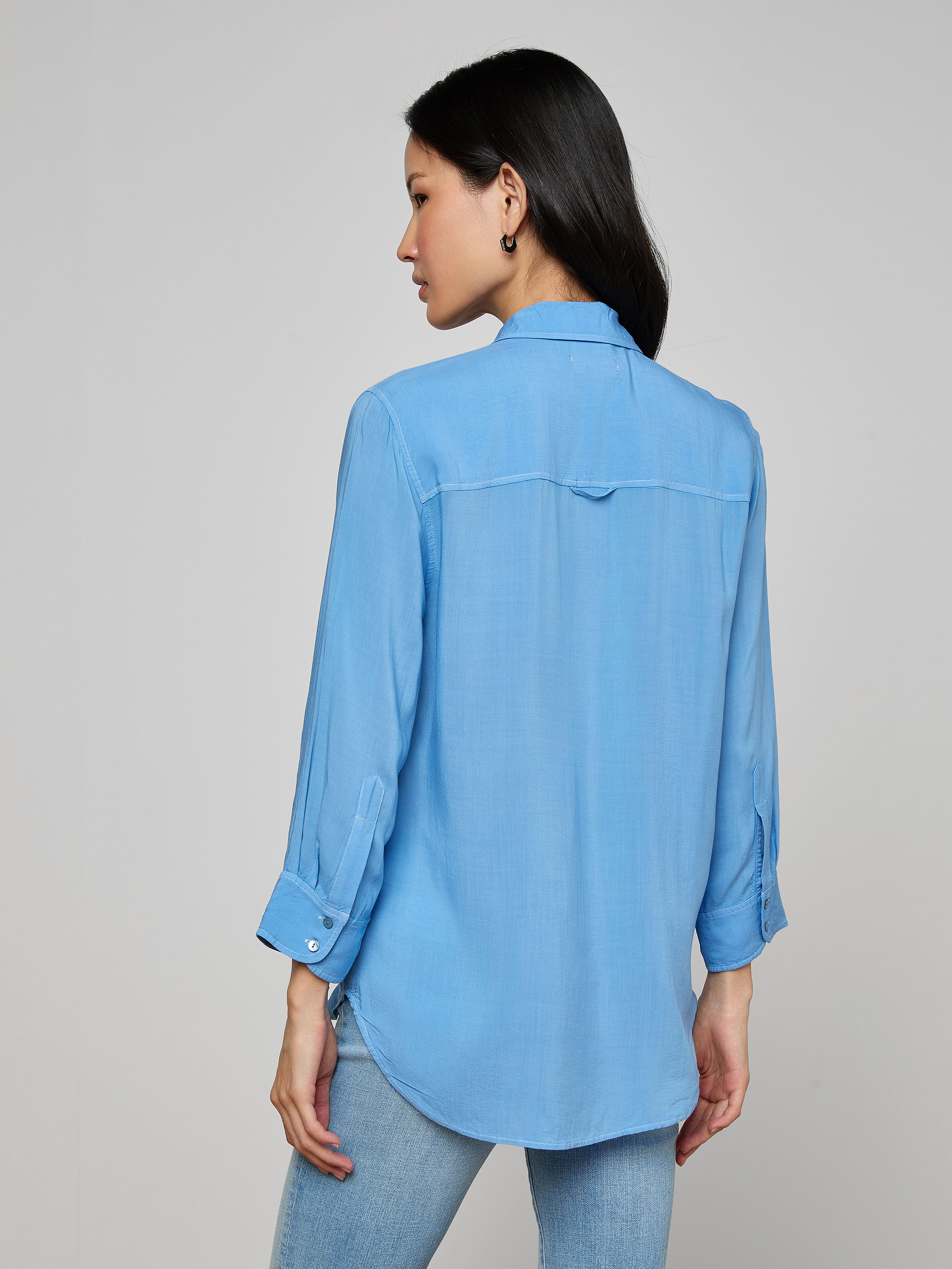 A woman stands facing away, wearing the Marlene Blouse—a lightweight button-down with rolled-up sleeves—paired with light blue jeans against a plain, light background, embodying everyday polish with its relaxed fit.