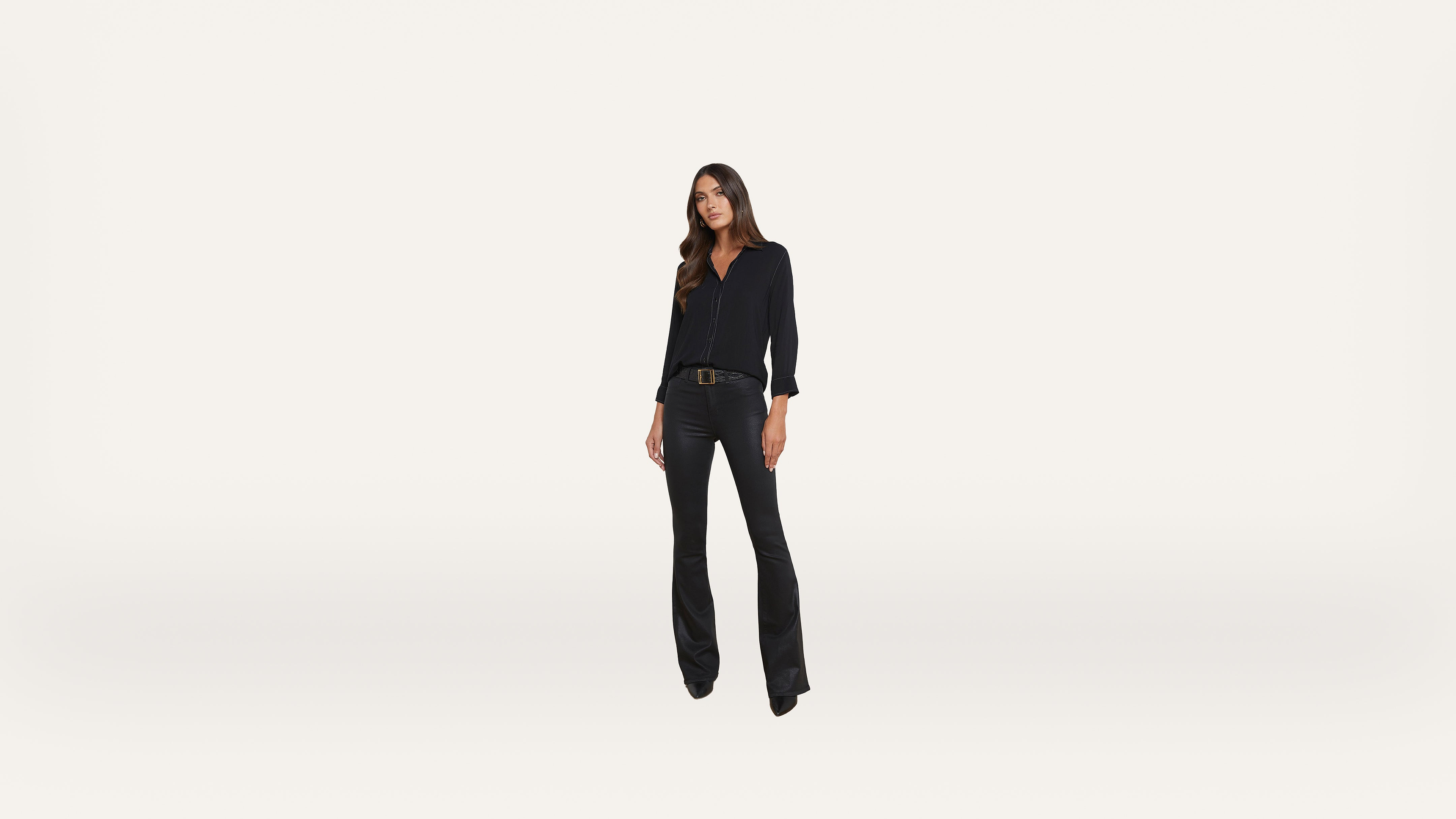 A woman with long hair stands facing forward, wearing a black long-sleeve top, black flared pants, and black shoes against a plain white background.