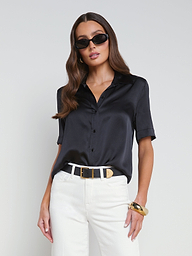 Wearing the Ellah Silk Blouse in black, paired with white pants and a black belt with a gold buckle, this woman accessorizes her look with sunglasses and gold jewelry as she stands against a plain light background—capturing effortless summer style.