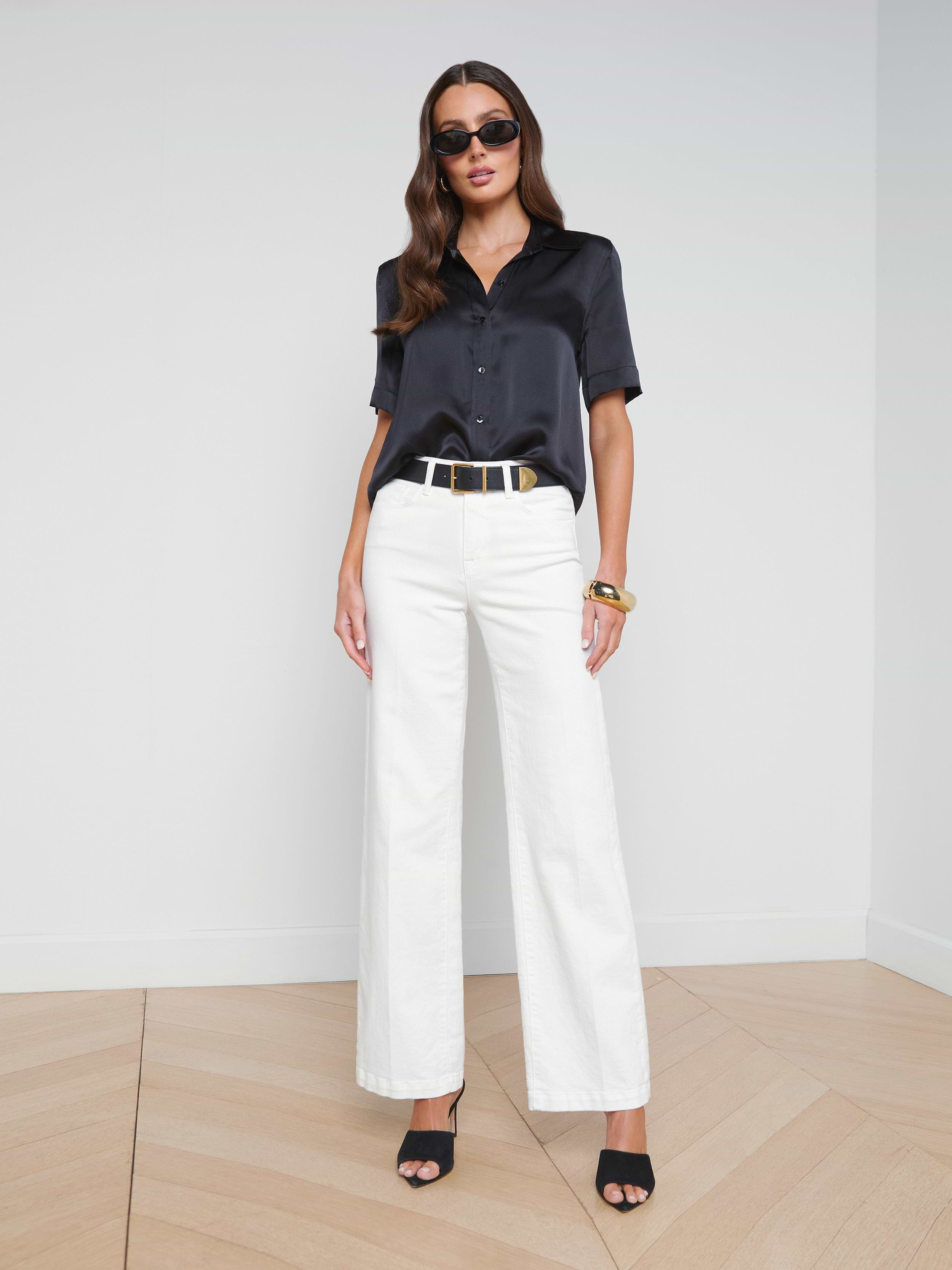 Wearing the Ellah Silk Blouse styled for a chic summer look, a woman pairs it with white wide-leg pants, black heels, a black belt, and sunglasses as she stands against a plain white background.