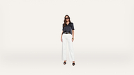 A woman wearing sunglasses, a dark short-sleeve shirt, white wide-leg pants, and black heels stands against a plain white background.