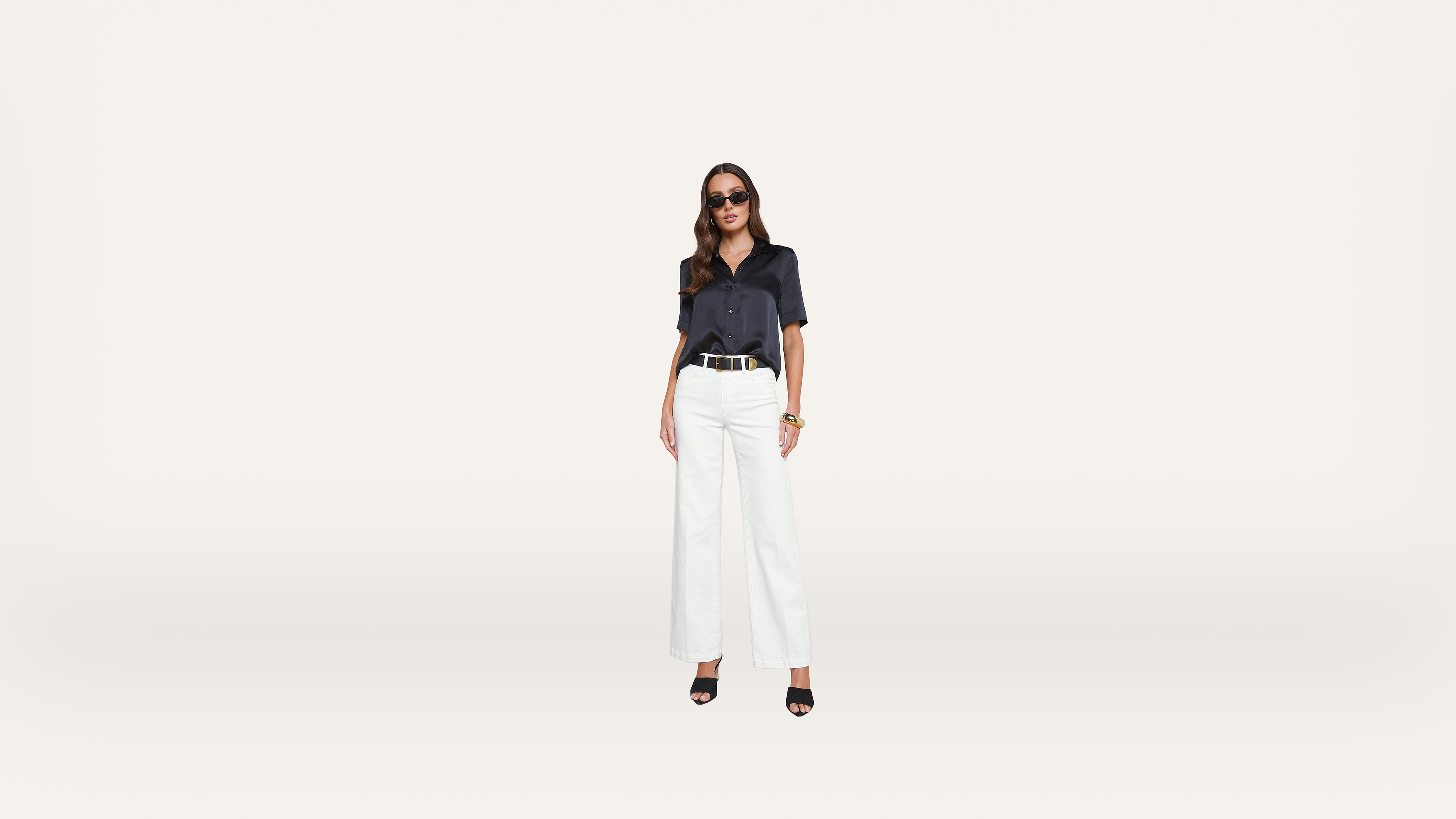 A woman wearing sunglasses, a dark short-sleeve shirt, white wide-leg pants, and black heels stands against a plain white background.