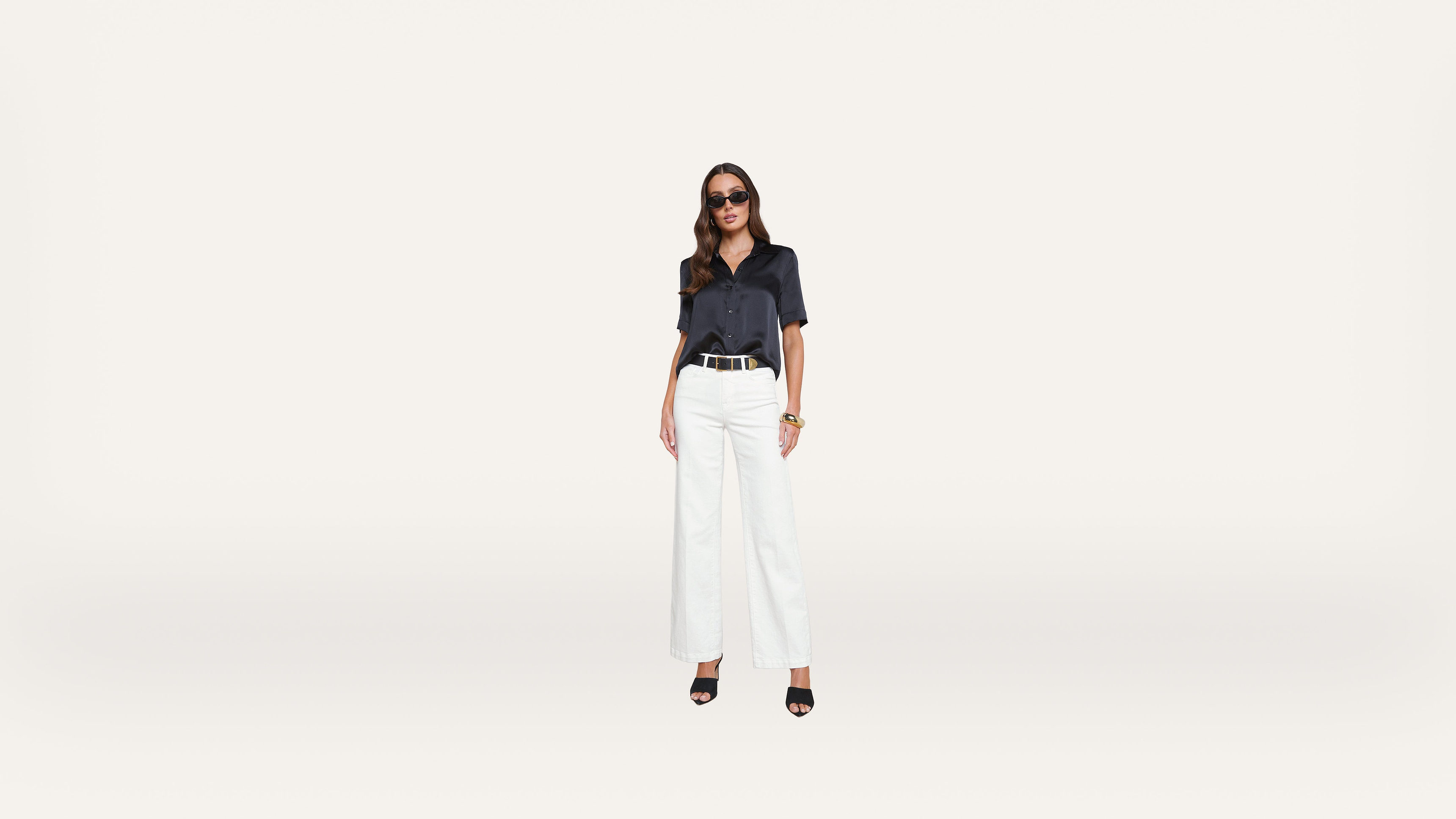 A woman wearing sunglasses, a dark short-sleeve shirt, white wide-leg pants, and black heels stands against a plain white background.