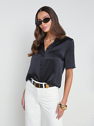 Wearing the Ellah Silk Blouse in black with short sleeves, white pants, dark sunglasses, a black and white belt, and a gold bangle, a woman stands against a plain light background, embodying effortless summer style.