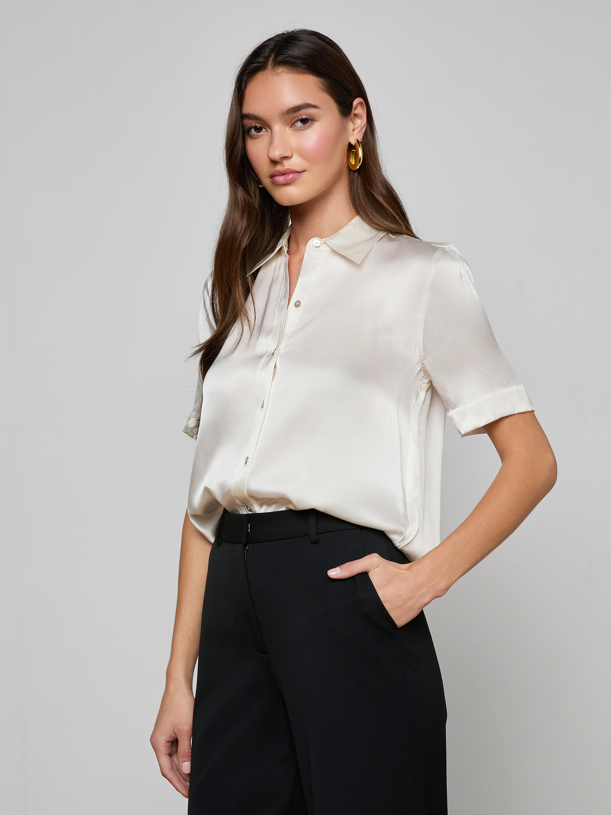 Wearing the Ellah Silk Blouse in white, a woman with long brown hair pairs it with white pants, a beige belt featuring a gold buckle, and gold earrings as she stands against a plain light background.