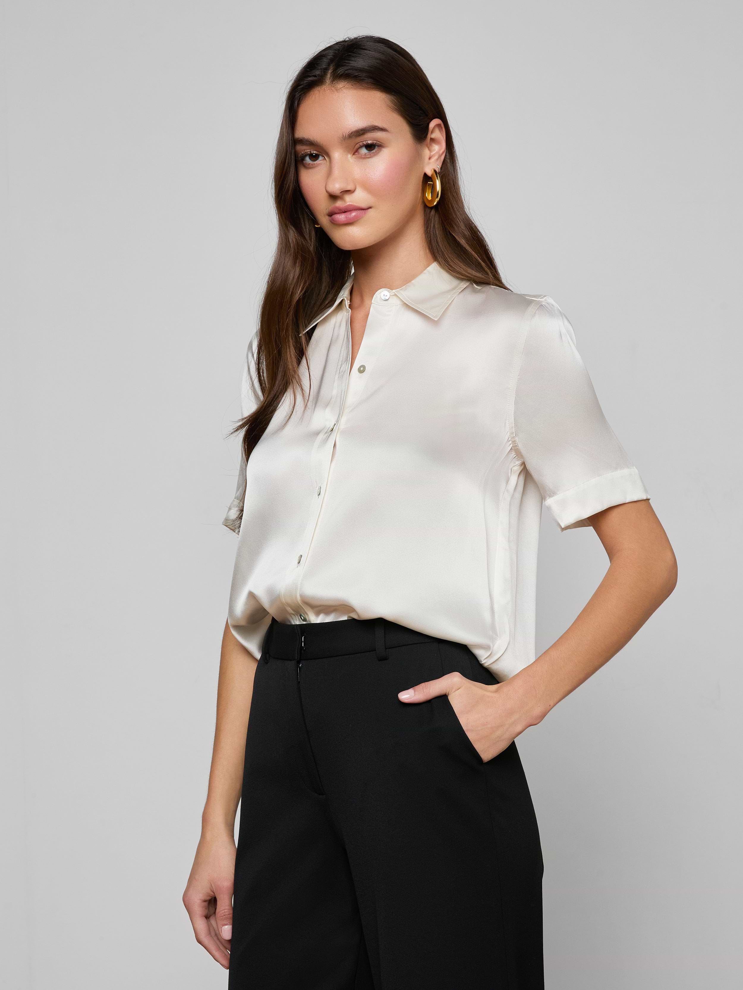 Wearing the Ellah Silk Blouse in white, a woman with long brown hair pairs it with white pants, a beige belt featuring a gold buckle, and gold earrings as she stands against a plain light background.