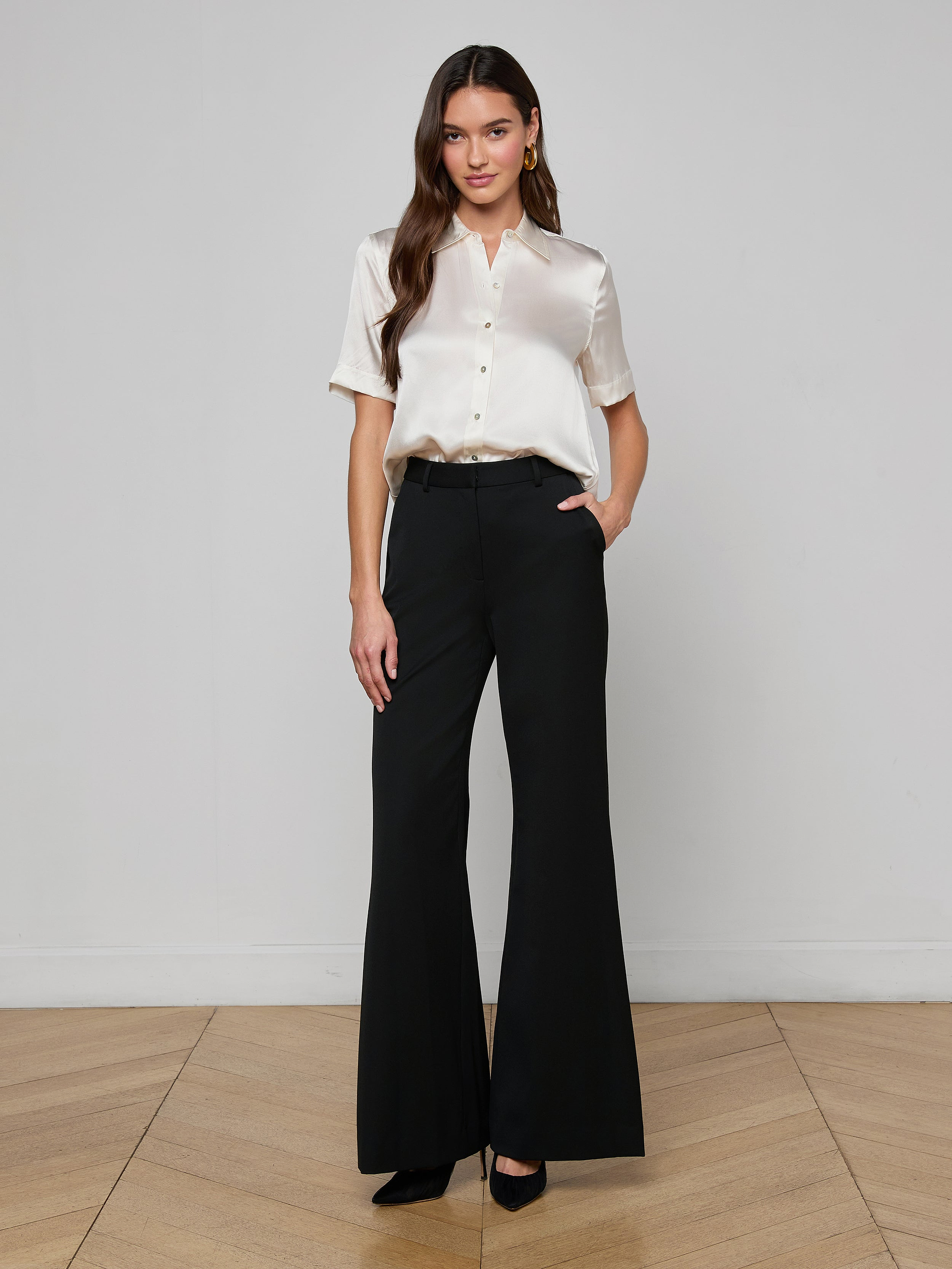 A woman stands against a plain white background, wearing the Ellah Silk Blouse with short sleeves, cream pants, a belt, and brown high heels. She has long brown hair and a neutral expression.