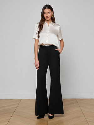 A woman stands against a plain background, smiling slightly, wearing the Ellah Silk Blouse with short sleeves in cream, paired with high-waisted black wide-leg pants and black heels. One hand rests in her pocket.
