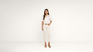 A woman stands against a plain white background, wearing a sheer white blouse, beige cropped pants, a belt, and brown heels. One hand is in her pocket, and she looks directly at the camera.