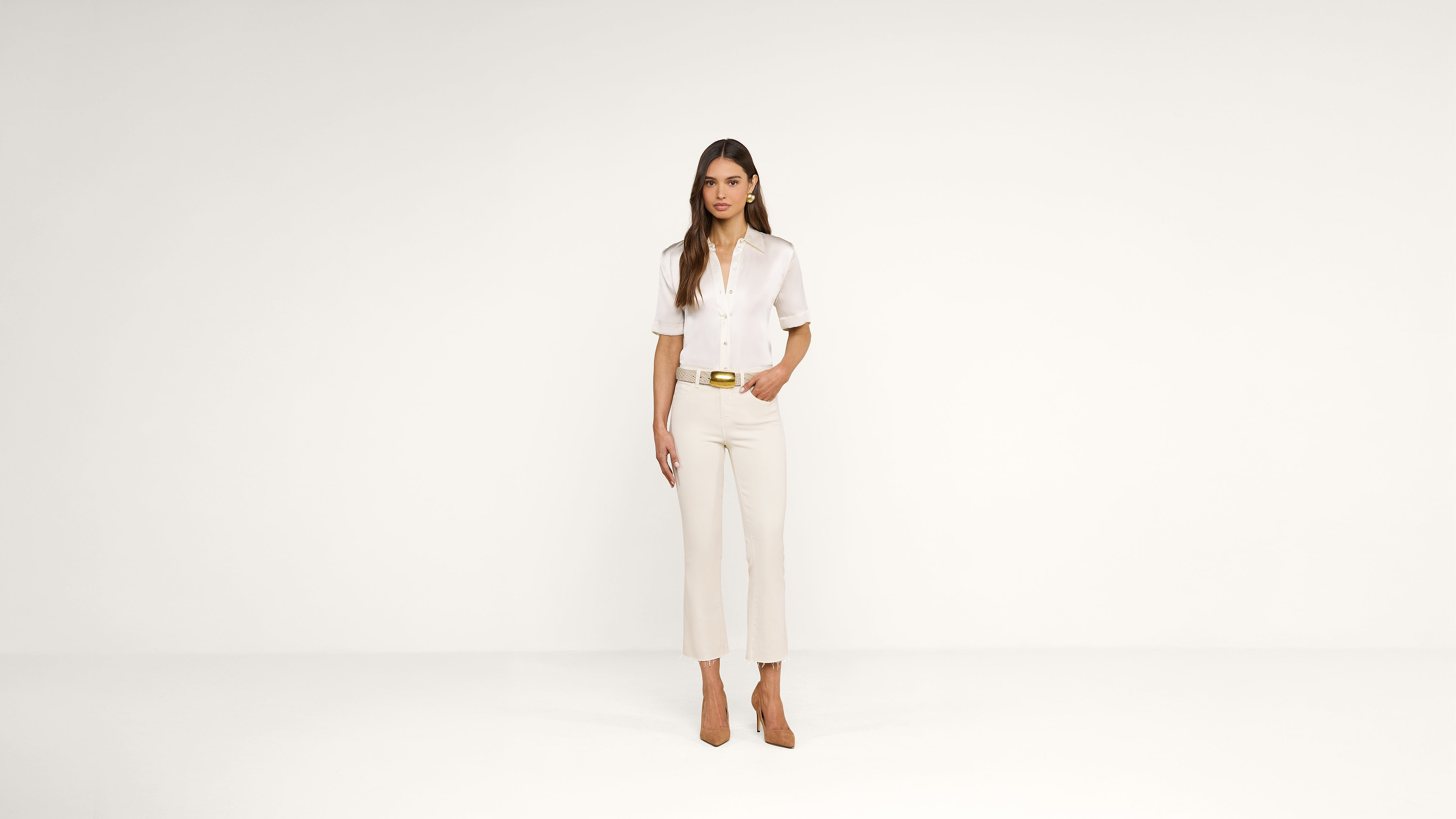 A woman stands against a plain white background, wearing a sheer white blouse, beige cropped pants, a belt, and brown heels. One hand is in her pocket, and she looks directly at the camera.