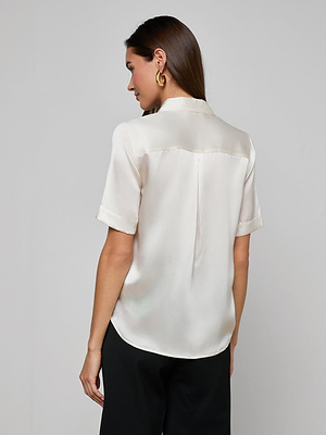 A woman with straight brown hair faces away, wearing the Ellah Silk Blouse in ivory with short sleeves, paired with black pants and gold hoop earrings, posed against a plain light gray background.