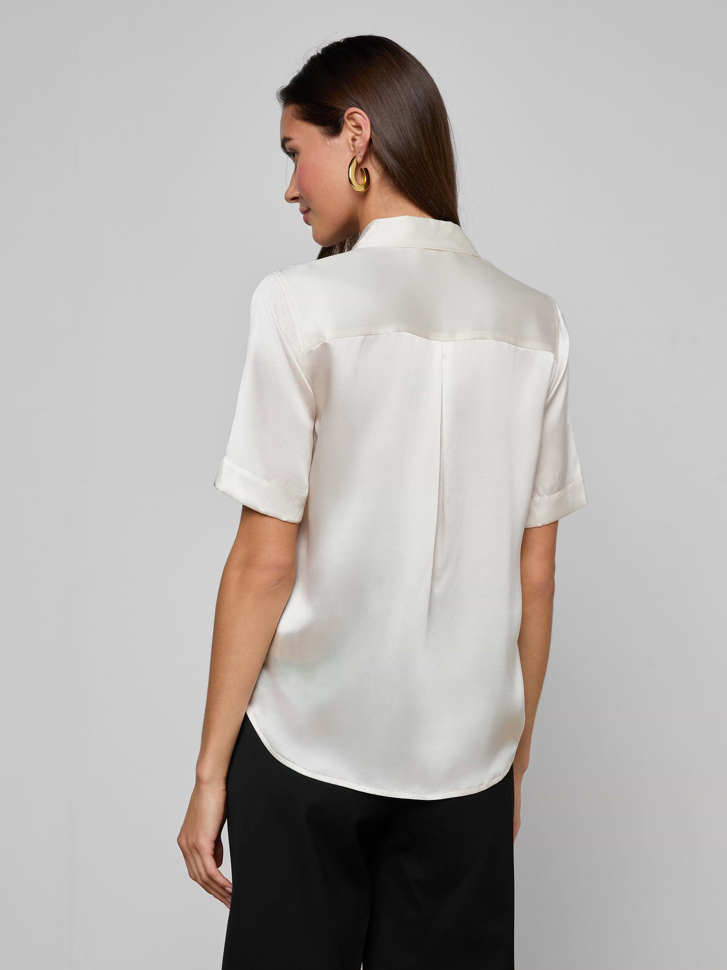 A woman stands facing away, wearing the Ellah Silk Blouse in a short-sleeved, cream-colored silk charmeuse along with matching pants against a plain light background.