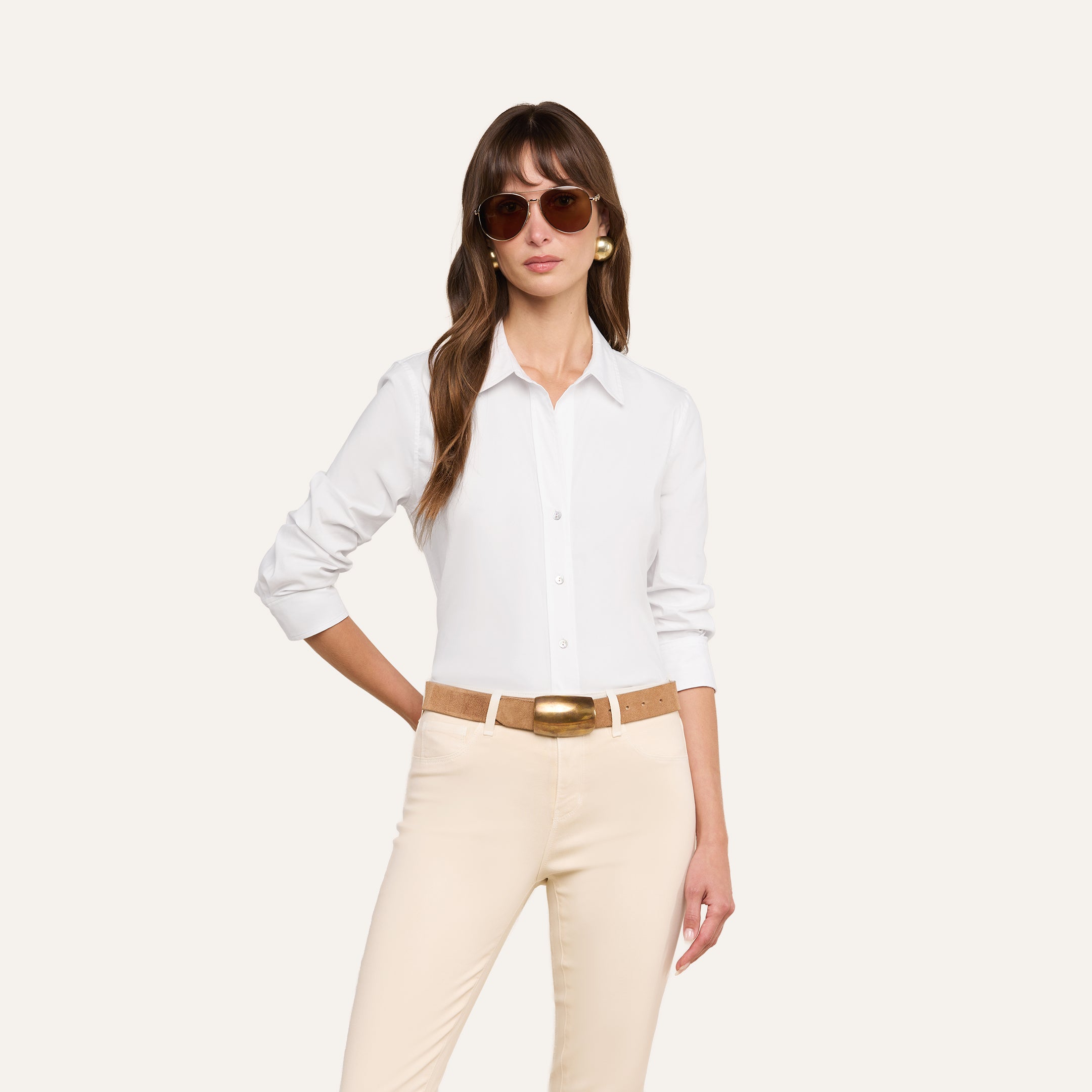A woman wearing sunglasses and the Daniella Cotton-Blend Blouse—a white, stretch cotton poplin top with three-quarter sleeves—paired with beige pants and a tan belt featuring a large gold buckle, stands with one hand on her hip against a plain light background.