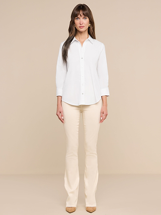 A woman stands facing forward against a beige backdrop, wearing the Daniella Cotton-Blend Blouse in white, cream pants, and tan shoes. Her long brown hair frames her face as her hands rest by her sides.