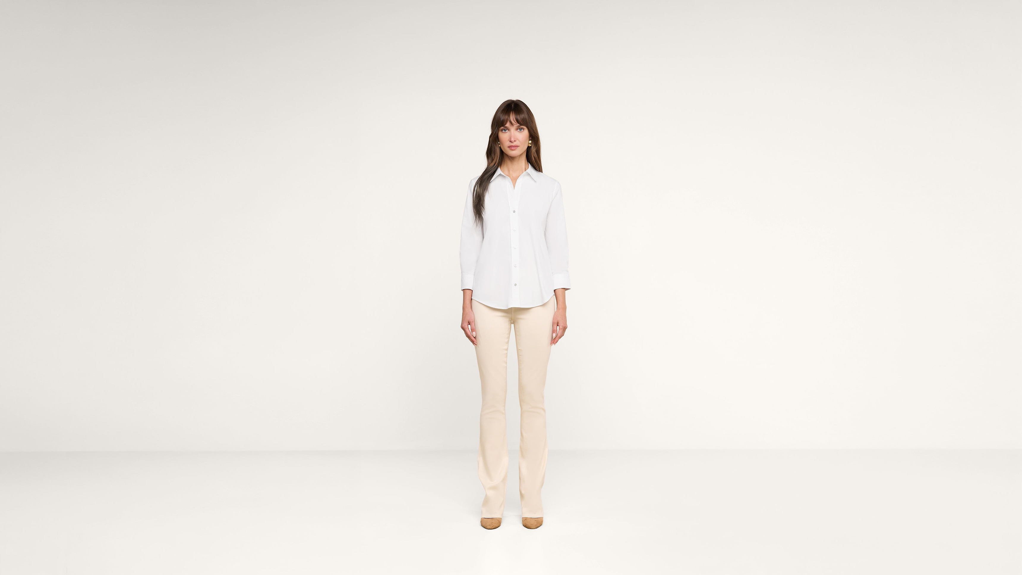 A woman with long brown hair stands in a minimalist, white room. She wears a white button-up shirt, beige pants, and tan shoes, facing forward with a neutral expression.