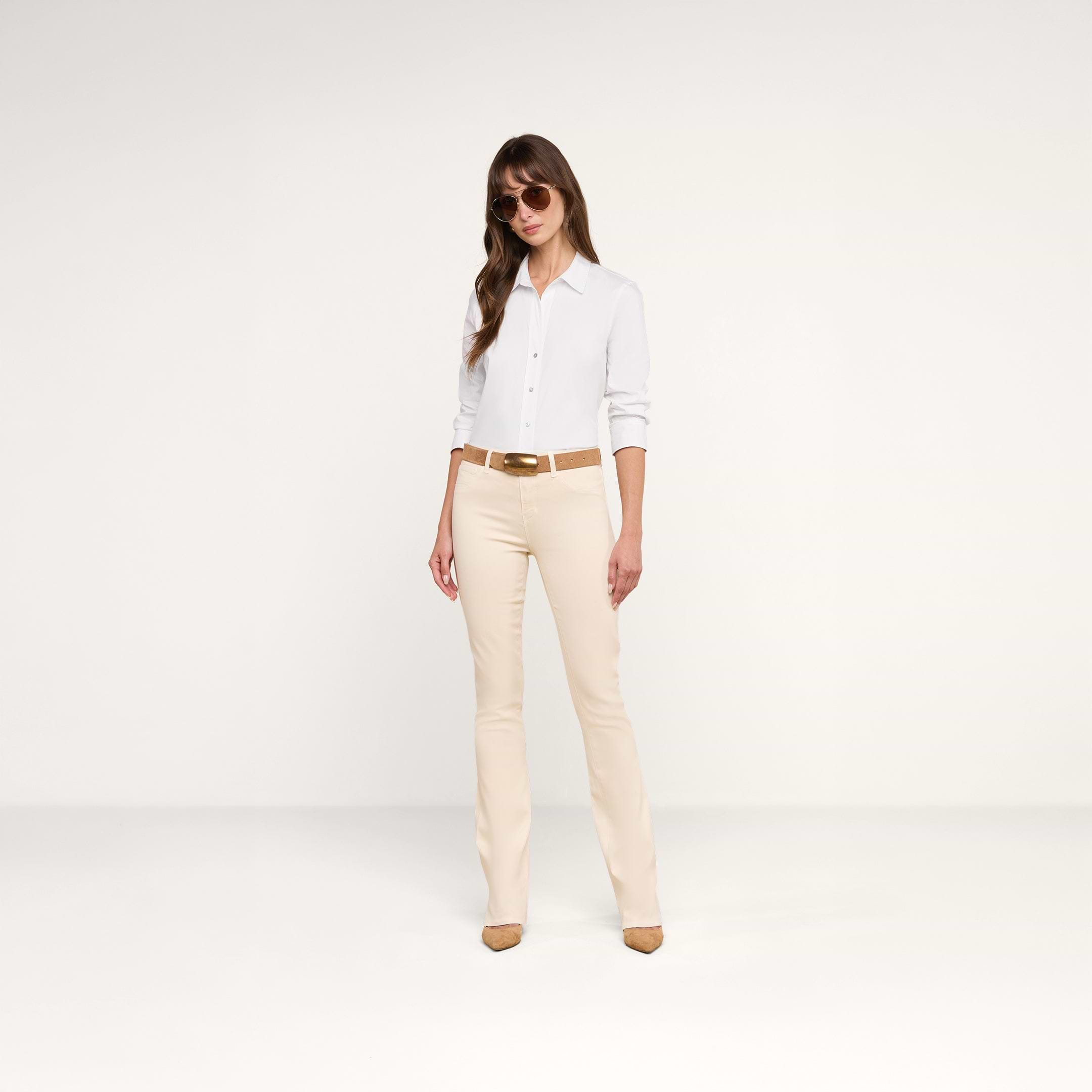 A woman wearing sunglasses, the Daniella Cotton-Blend Blouse with three-quarter sleeves, beige pants with a brown belt, and beige heels stands against a plain white background.