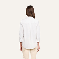 A person with long brown hair, seen from behind, wears the Daniella Cotton-Blend Blouse— a relaxed-fit white stretch cotton poplin top with three-quarter sleeves—paired with cream pants, standing against a plain light background.